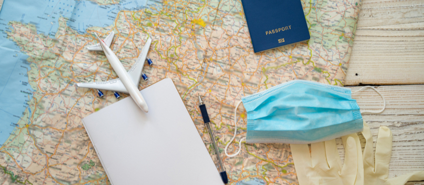 A world map with a figurine plane and passport and pen and notepad with medical gloves and masks on it