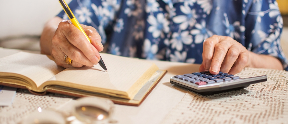 Person writing down in notepad with calculator and paperwork to the side