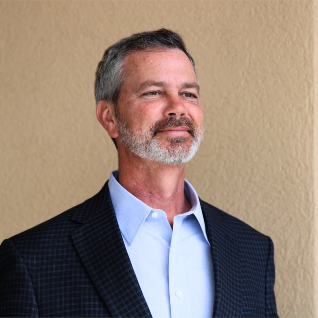 Man stands against a tan stucco wall wearing a blue blazer and a light blue collard shirt.