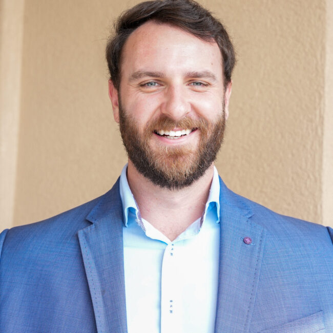Man stands in front of a tan stucco wall wearing a blue blazer and a light blue collared shirt