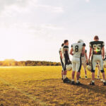 A football team huddled up on a field with the sun shinning behind them.