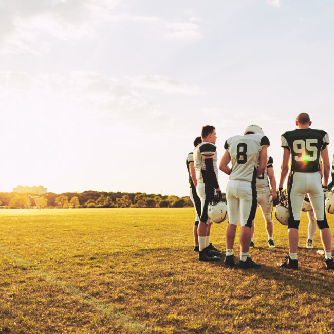 A football team huddled up on a field with the sun shinning behind them.